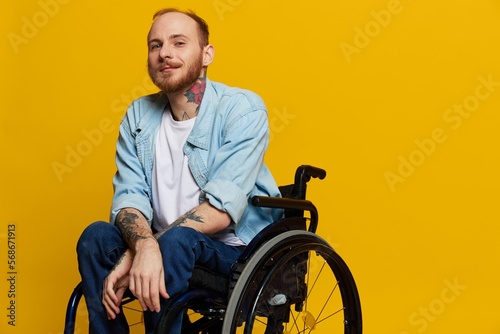 A man in a wheelchair smile and happiness, thumb up, with tattoos on his hands sits on a yellow studio background, the concept of health a person with disabilities