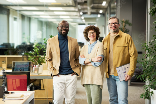 Three successful managers or brokers in smart casualwear looking at camera while standing in openspace office with green plants