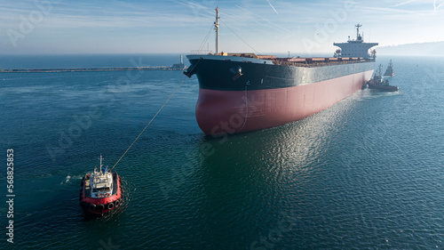 Aerial view of tug boat assisting big cargo ship. Large cargo ship enters the port escorted by tugboats.