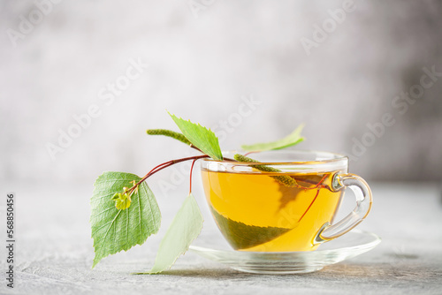 Glass cup of hot, fragrant birch tea on a grey background and a branch of birch leaves