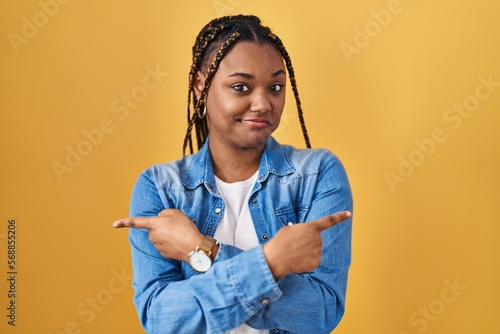 African american woman with braids standing over yellow background pointing to both sides with fingers, different direction disagree