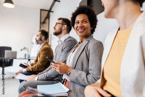 A multiracial mentees are sitting at corporate firm and waiting for the meeting at the office. The african woman is using phone while chatting with colleague.