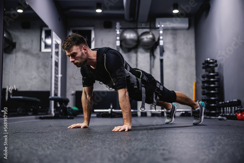 Modern training concept on electrical muscle stimulation. Side view of a male wearing an EMS suit and doing arm exercises and push-ups. He is in a plank position and he touching the floor with hands