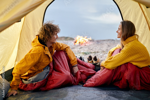 Beautiful couple sit inside of tent talking, young caucasian man and woman in love, in nature, in sportive clothes, bonfire in the background, in the evening. camping, travel, adventure concept