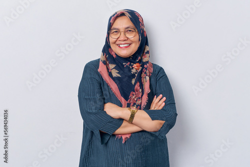 Elderly smiling Asian Muslim woman 50s wearing hijab in glasses crossed arms and looking confident isolated on white background. People islam religious concept