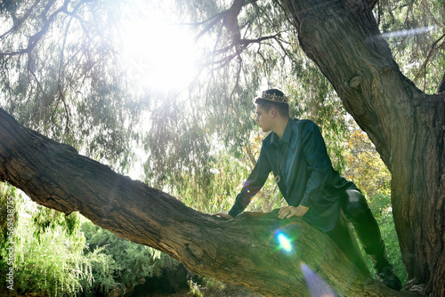 portrait of handsome masked man wearing fantasy medieval prince costume with golden crown and romantic silk shirt. climbing a tree branch in a forest location with backlit silhouette lighting.