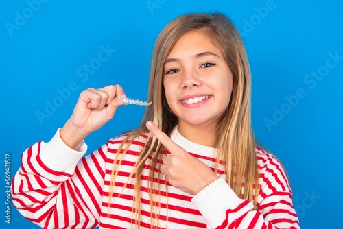 caucasian teen girl wearing striped shirt over blue studio background holding an invisible aligner and pointing at it. Dental healthcare and confidence concept.