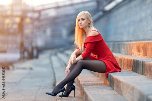 Beautiful blonde girl in a red dress with perfect legs in pantyhose and shoes with high heels posing outdoor on the city square.