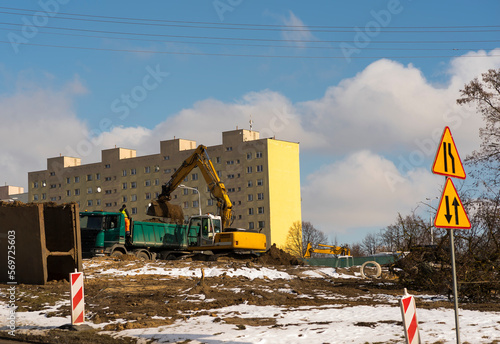 Plac budowy . Duża ,gąsienicowa koparka ładuje ziemię na ciężarówkę . Na tle bloku mieszkalnego i błękitnego nieba z chmurkami . 