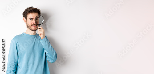 Smiling young man detective looking through magnifying glass, searching for promo offer, standing on white background