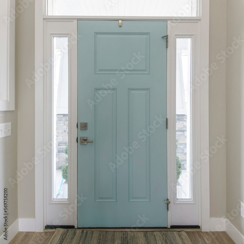 Square Mint front door interior with transom window and sidelights. Interior of a house with light gray walls, wooden flooring and a view of two rooms on both sides with windows.