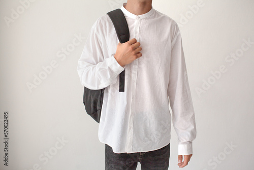 The guy with a bagpack on his shoulders is poaing on a white background. The teenager is wearing a white T-shirt and black jeans