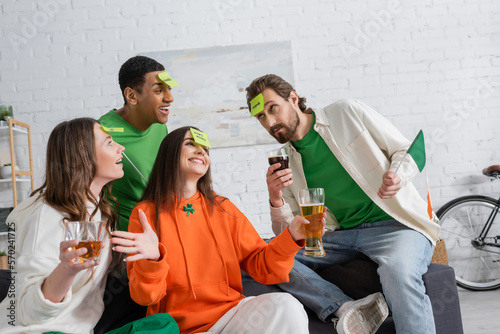 cheerful multiethnic friends with sticky notes on foreheads holding alcohol drinks and playing guess who game on Saint Patrick Day