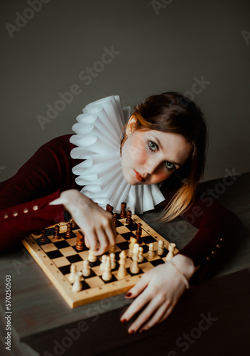 A beautiful woman in burgundy clothes and a white jabot sits at a table and plays wooden chess. The Queen's gambit. Mind and logic. Jester.