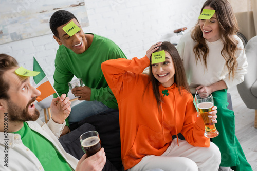 joyful multiethnic friends with sticky notes on foreheads holding alcohol drinks and playing guess who game on Saint Patrick Day