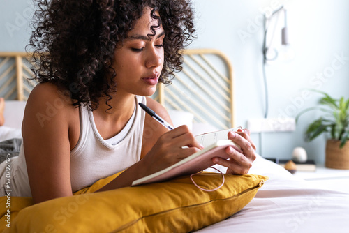 Pensive young multiracial latina woman lying down on bed with serious expression writing on journal in cozy bedroom