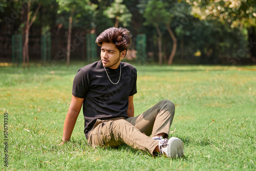 Attractive young indian man portrait in black t shirt and silver neck chain sitting on green lawn in public park, hindu male portrait. Handsome indian man portrait with thick hair in city park