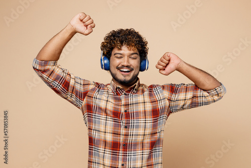 Young Indian man wear brown shirt casual clothes headphones listen to music dance on party have fun gesticulating hands isolated on plain pastel light beige background studio People lifestyle concept
