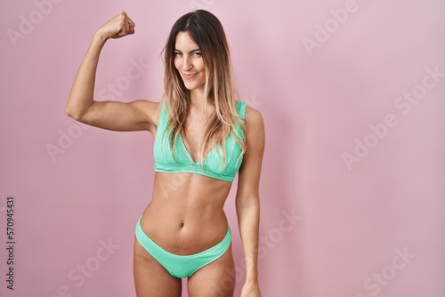 Young hispanic woman wearing bikini over pink background strong person showing arm muscle, confident and proud of power