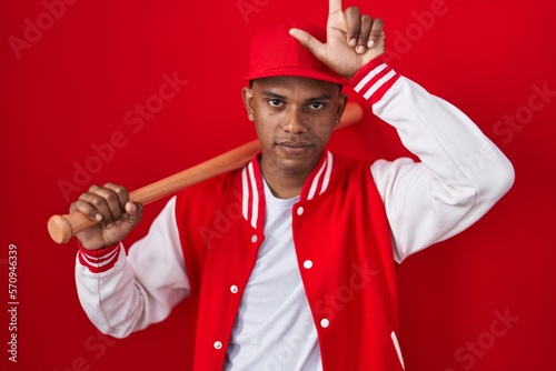 Young hispanic man playing baseball holding bat making fun of people with fingers on forehead doing loser gesture mocking and insulting.