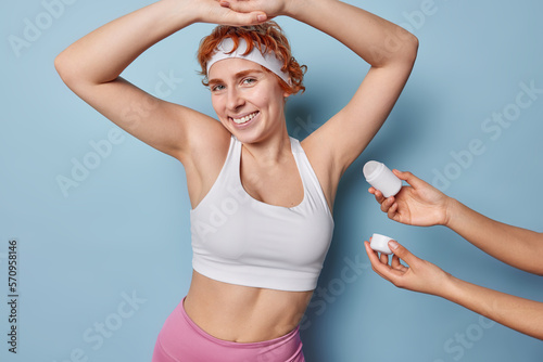 Horizontal shot of redhead sporty woman stands satisfied keeps arms up smiles toothily dressed in cropped top has fit body uses deodorant roll antiperspirant bottle isolated over blue background.