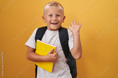 Little caucasian boy wearing student backpack and holding book celebrating victory with happy smile and winner expression with raised hands