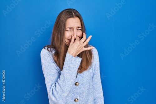 Young woman standing over blue background smelling something stinky and disgusting, intolerable smell, holding breath with fingers on nose. bad smell