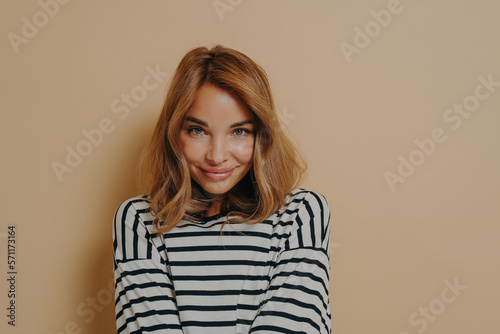 Young charming woman with dyed blonde hair in striped shirt looking at camera with shy smile