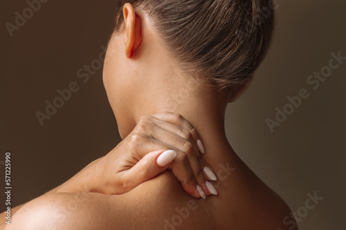 Cropped shot of a young woman standing with her back holds her neck massaging a trapezius muscle with her hand isolated on dark brown background. The girl suffers from an osteochondrosis