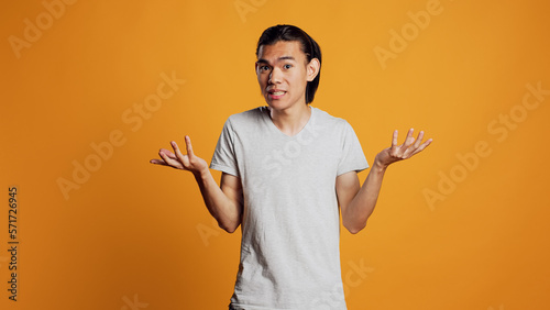 Clueless young person doing i dont know sign in studio, acting uncertain and unsure about answer to question. Asian guy being carefree and casual, acting doubtful over orange backdrop.