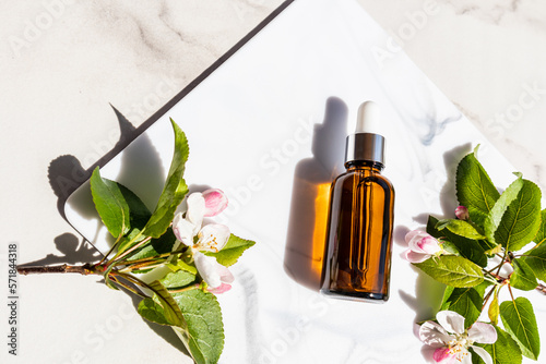 a cosmetic bottle of dark glass with a dropper lies on a white marble podium with flowering branches of an apple tree. top view. natural cosmetics.