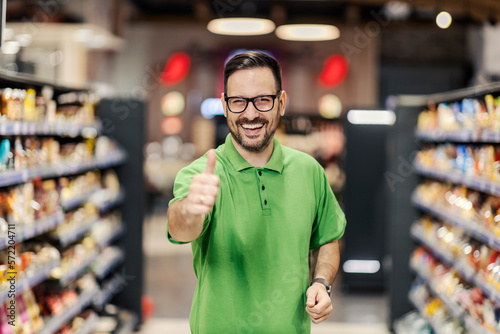 A happy employee is standing at the supermarket between the aisles and giving thumbs up at the camera.