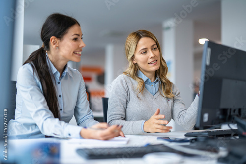 Two female business colleagues working together and discussing project on computer while sitting at desk in office.