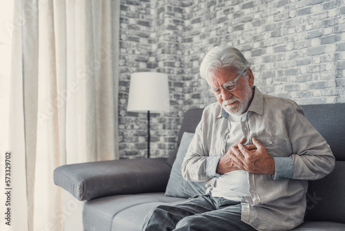 Worried elder senior man feeling bad, upset old middle aged grandfather touching chest feel sudden pain heartburn having heart attack sit on sofa at home.