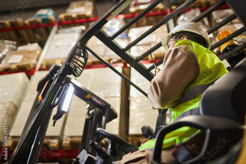 Low angle view of female worker wearing hardhat while operating forklift truck in warehouse and talking to portable radio