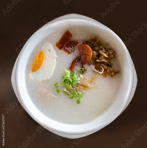 High angle view, rice porridge with salted boil egg, Thai sausage, minced pork and spring oonion on top in white bowl. Asian breakfast at the hotel, hot cuisine concept. Delicious congee at restaurant