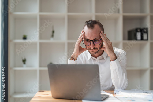 Young unhappy man office worker feeling bored at work, looking at laptop with demotivated face expression while sitting at workplace in office, distracted male worker feeling tired of monotonous job
