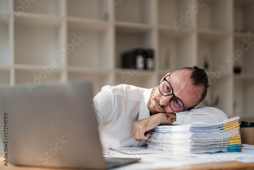 Young unhappy man office worker feeling bored at work, looking at laptop with demotivated face expression while sitting at workplace in office, distracted male worker feeling tired of monotonous job