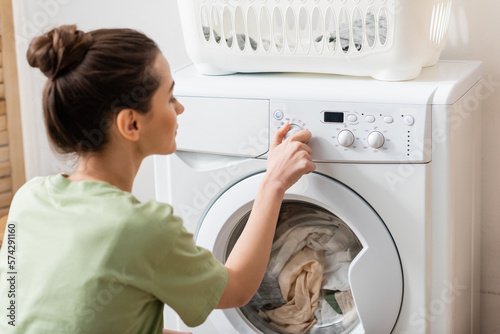 Side view of blurred woman switching washing machine near basket in laundry room.