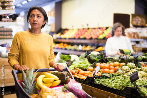 Pensive latina buying groceries at store, walking with shopping cart among shelves and choosing products