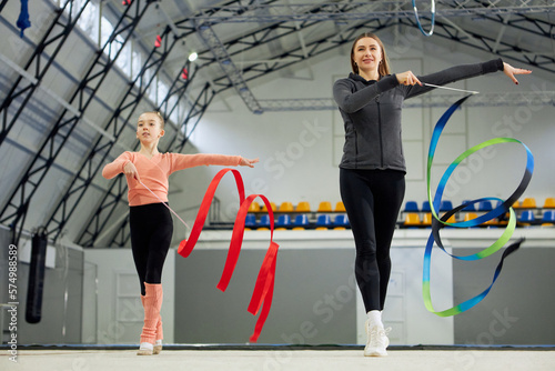 Little girl, child, rhythmic gymnast training indoors with coach, doing exercise with ribbon. Concept of sportive lifestyle, childhood, education, health, professional sport, championship