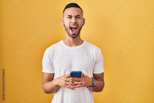 Young hispanic man using smartphone typing message angry and mad screaming frustrated and furious, shouting with anger looking up.