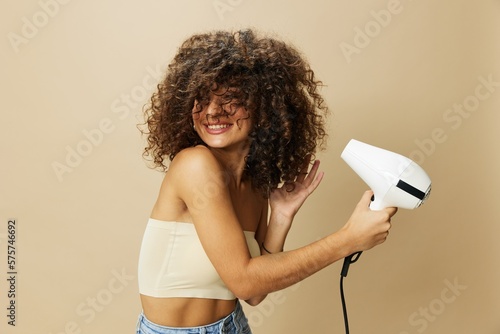 Woman dries curly afro hair with blow dryer, home beauty care styling products hair, smile on beige background