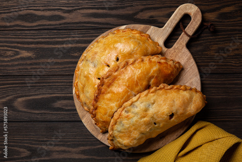 Flat la of baked meat turnovers or pies, or empanadas, or cornish pasty with filling, beef, carrot, and potato on a dark wooden board.