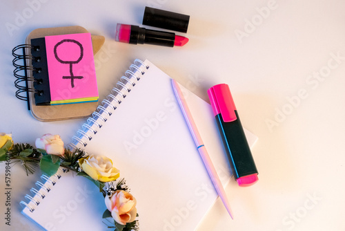 Material de oficina con una corona de flores sobre la libreta, un pintalabios rosa y unos post it con el símbolo de genero femenino por el día internacional de la mujer.