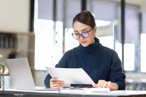 Young Asian professional business woman office worker analyst sitting at desk working on laptop thinking on project plan, analyzing marketing or financial data online, watching elearning webinar.