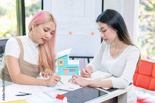 Concept of engineering consulting, Two female engineers discussing about model of building together; Two female architects are studying blueprint of building house.