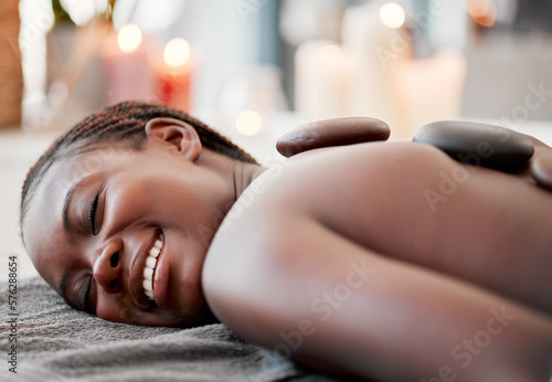 Hot stone massage, black woman at spa with smile on face, holistic healing with physical therapy and back skin detox. Happy, content and zen with wellness at luxury resort, rocks and relax with glow