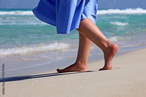 Barefoot woman in blue dress walking by the sand on sea waves background. Female legs on a coast, beach vacation