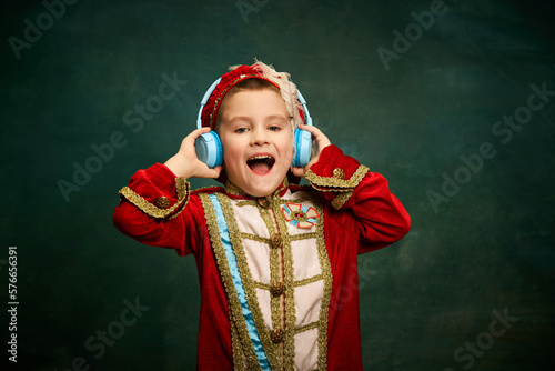 Cheerful little boy dressed up as medieval little prince and pageboy listening to music over dark vintage style background. Retro fashion, emotions, music concept